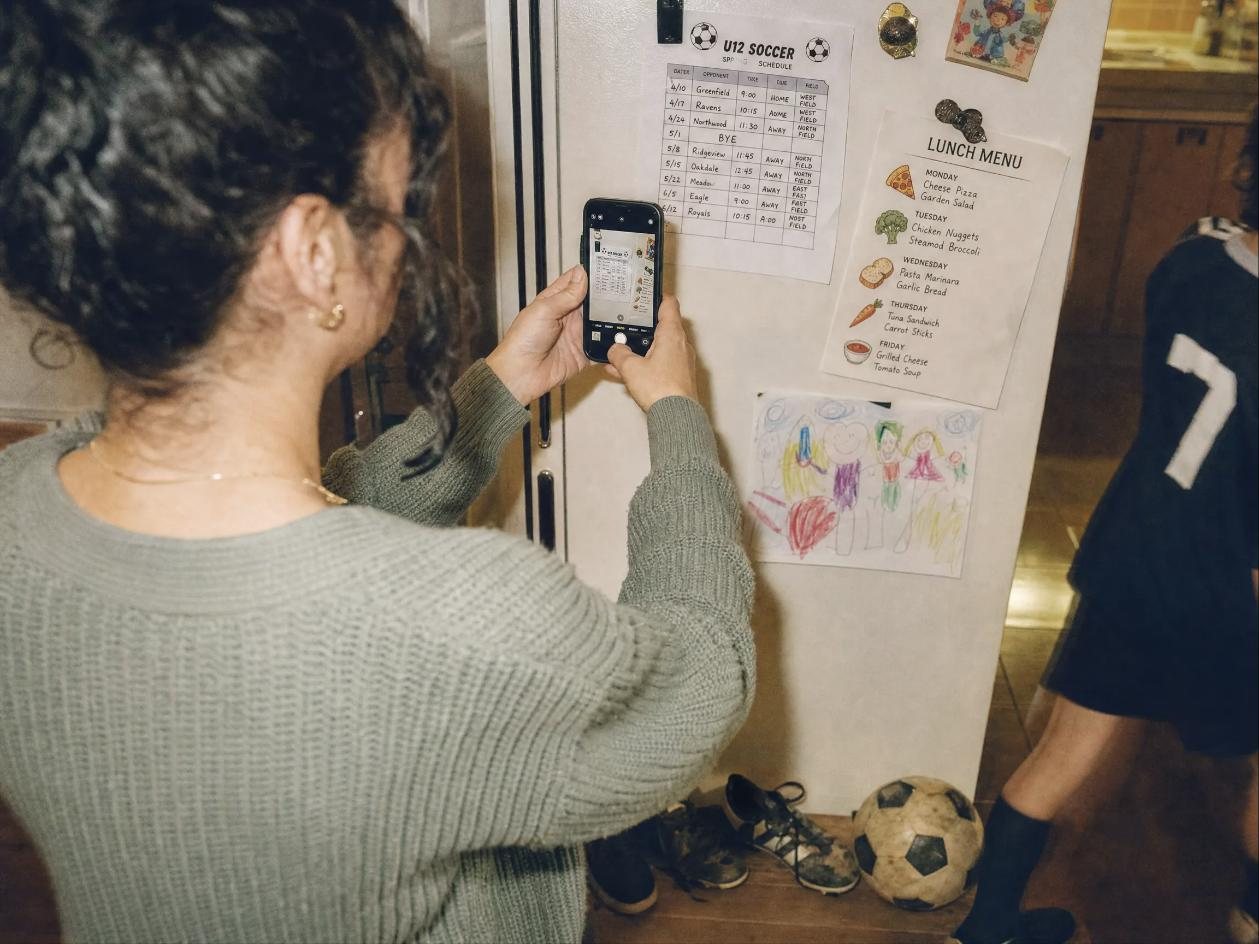 A mom photographs a sports schedule on the fridge with her phone